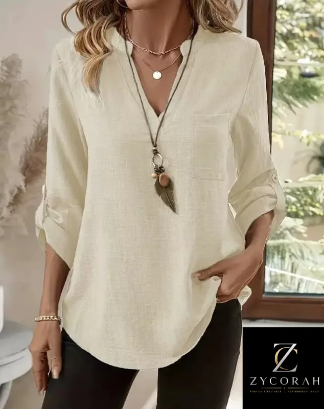 A woman wearing a loose, off-white linen long-sleeve blouse with a chest pocket and rolled sleeves, paired with black pants. The photo is taken in a softly lit indoor setting with a window in the background, and a 'ZYCORAH' brand logo is visible in the bottom right corner.