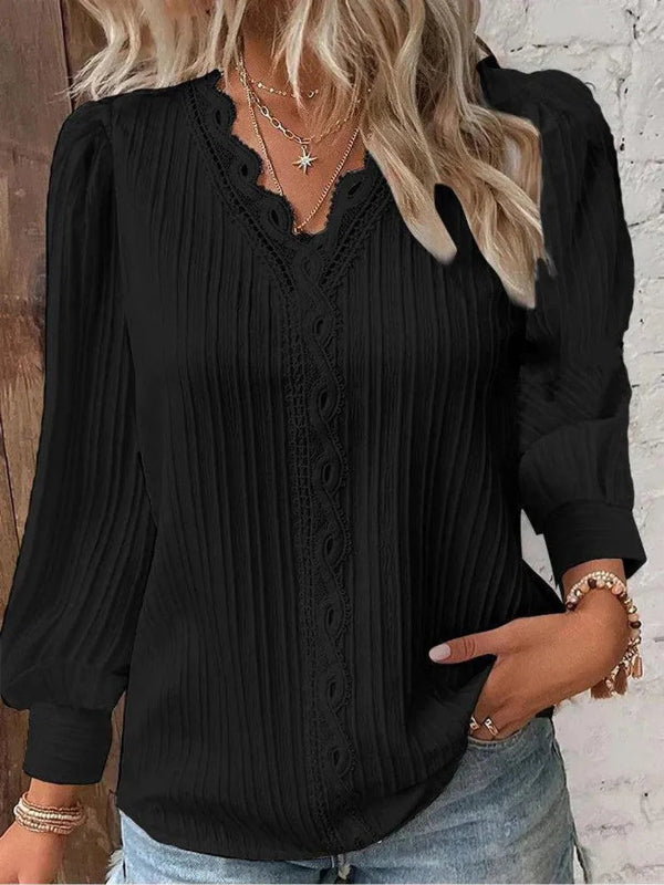 Black blouse with long sleeves and a V-neckline worn by a person, against a white brick wall.