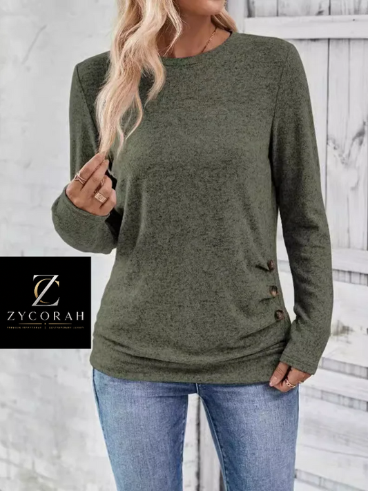 A woman wearing an olive green long-sleeve knit sweater with decorative buttons on the side, paired with blue jeans. A black brand overlay for 'Zycorah' is visible in the lower-left corner.