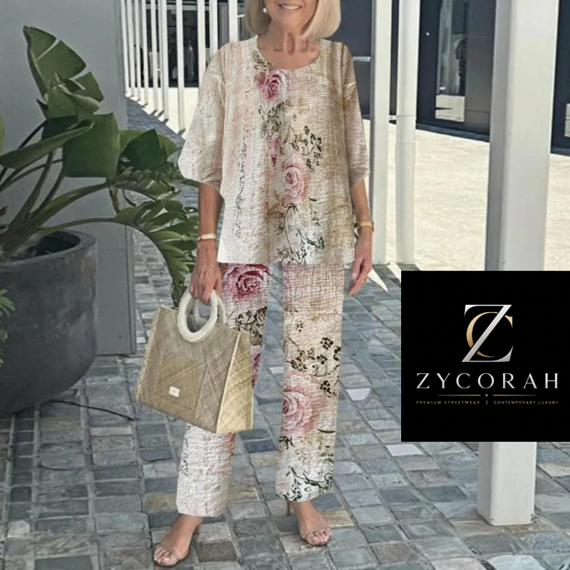 A woman models a two-piece matching lounge set from ZYCORAH, featuring a short-sleeve top and wide-leg pants in a beige linen-look fabric with pink and green floral prints. She is standing on a stone patio next to a potted plant, holding a woven handbag.