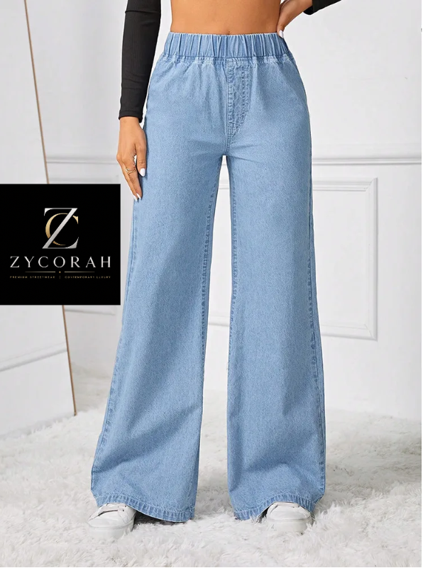 A model wears light blue wide-leg denim jeans with an elastic waistband, paired with a black long-sleeve top and white sneakers. A black brand card for 'Zycorah' is displayed on the left side against a white paneled wall and white rug background.