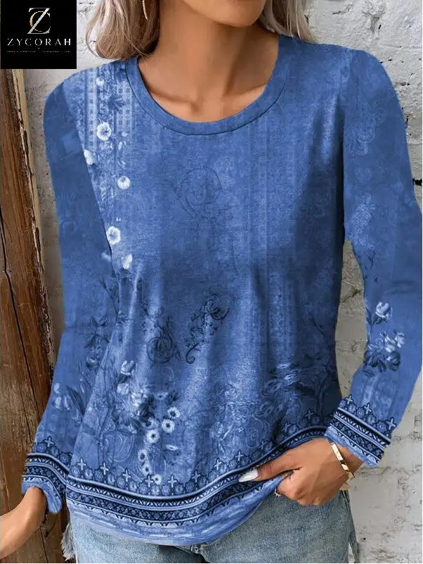 A woman wearing a blue long-sleeve top with a faded floral and paisley print, paired with light-wash jeans. The top features a round neckline and decorative patterned trim along the cuffs and hem.