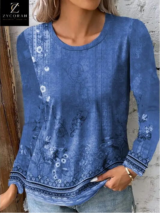 A woman wearing a blue long-sleeve top with a faded floral and paisley print, paired with light-wash jeans. The top features a round neckline and decorative patterned trim along the cuffs and hem.