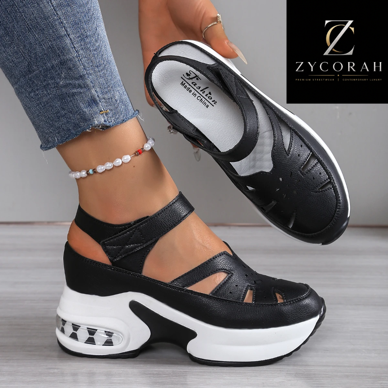 A person wearing a pair of black platform wedge sandals with cutout straps and white soles, shown against a neutral indoor floor and wall. One shoe is worn on the foot, and the other is held up to show the side and top.