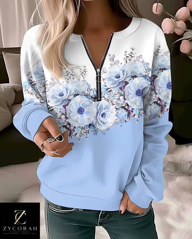 A woman wearing a light blue sweatshirt with a white upper section and a large floral print in blue, purple, and white across the chest. The sweatshirt has a half-zip neckline with a black zipper.