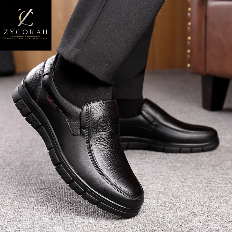 A pair of men's black slip-on leather shoes shown being worn with black dress pants, on a gray carpeted floor with a wooden chair in the background. The Zycorah brand logo is visible in the top left corner.