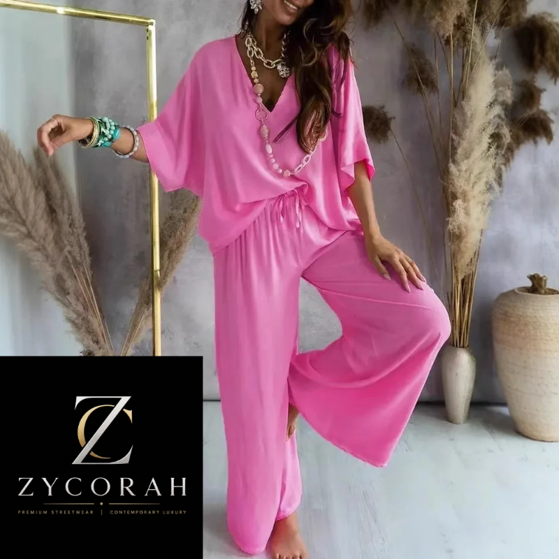 A woman models a bright pink two-piece lounge set consisting of a loose V-neck tunic top and matching wide-leg pants. The photo is styled in a modern interior with pampas grass, a gold mirror frame, and a ceramic vase, with a black brand overlay in the bottom-left corner reading 'Z YCORAH'.