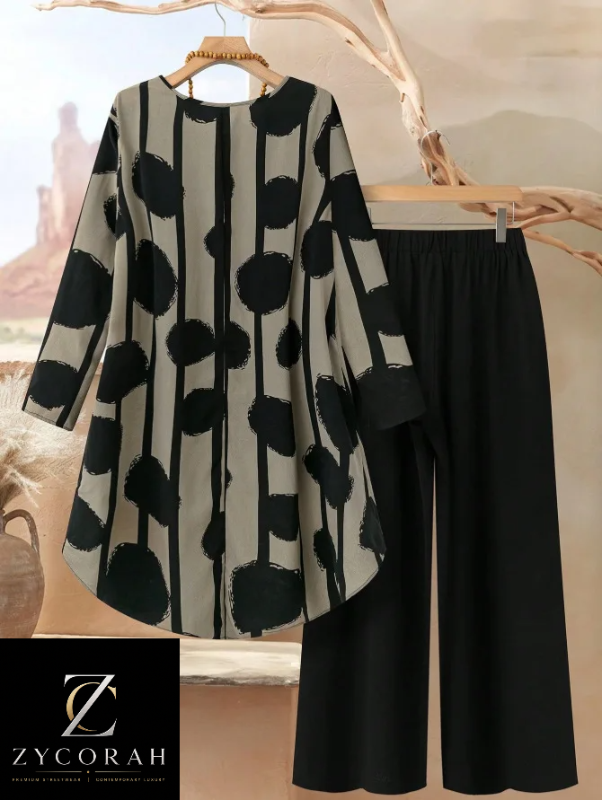 A women's two-piece outfit set featuring a long-sleeve tunic top with a black and beige abstract vertical stripe and polka dot pattern, paired with solid black wide-leg pants. The set is displayed on wooden hangers against a warm, rustic desert-themed backdrop with a Zycorah brand logo overlay in the bottom left corner.