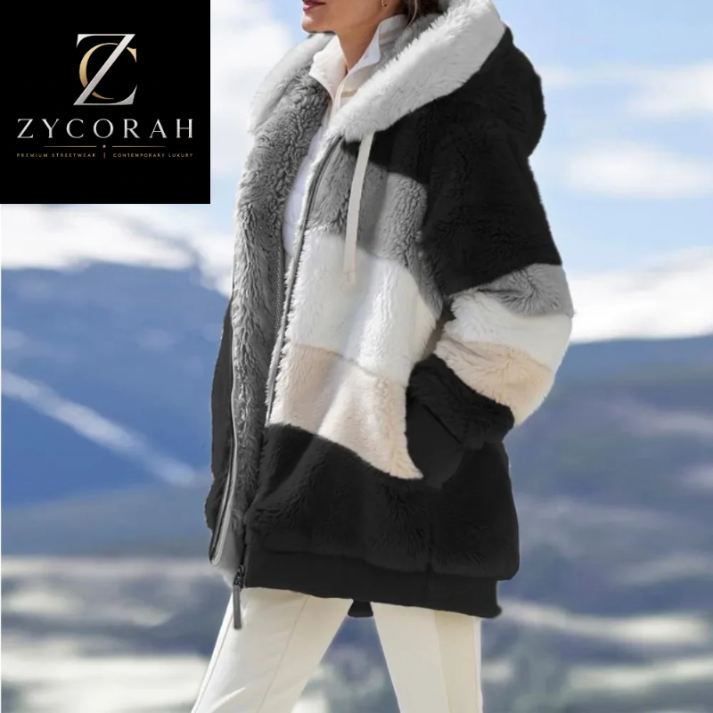 A woman wearing a plush color-blocked hooded jacket with black, white, gray, and beige panels, photographed outdoors against a mountain landscape. A black square logo in the top-left corner reads ZYCORAH.