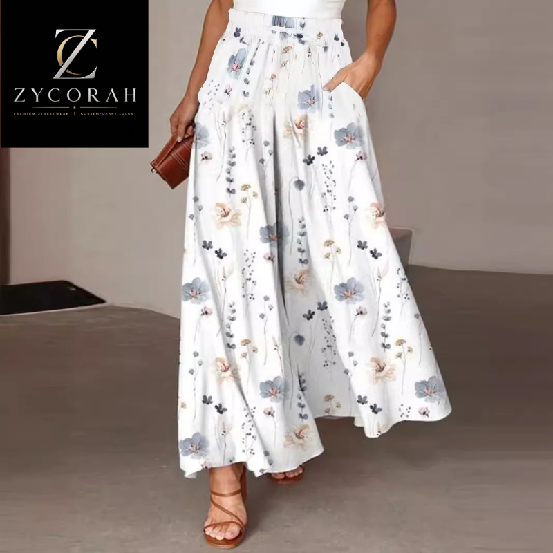 A model wearing white wide-leg maxi pants with a blue and beige floral print, paired with brown strappy sandals and holding a brown clutch. The Zycorah brand logo is visible in the top left corner.