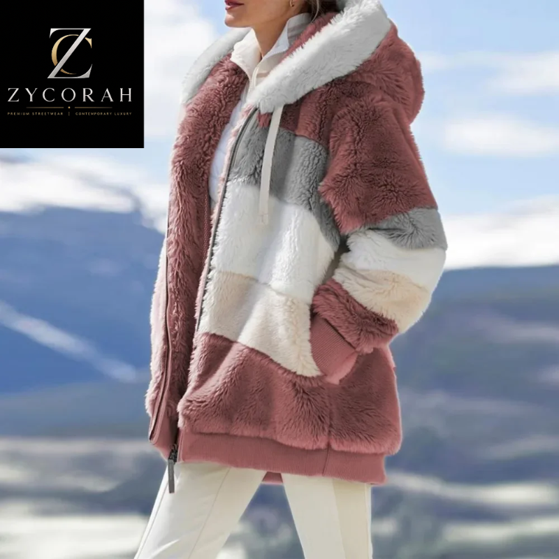 A woman wearing a plush faux-fur hooded jacket with large color-block panels in dusty pink, cream, white, and light gray, photographed outdoors against a blurred mountain landscape. A Zycorah logo appears in the top-left corner.