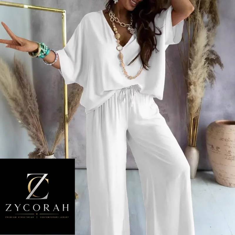 A woman models a white two-piece outfit consisting of a loose short-sleeve V-neck top and matching wide-leg drawstring pants. She is styled with layered gold necklaces and bracelets, standing in a studio setting with dried pampas grass and a ceramic vase.