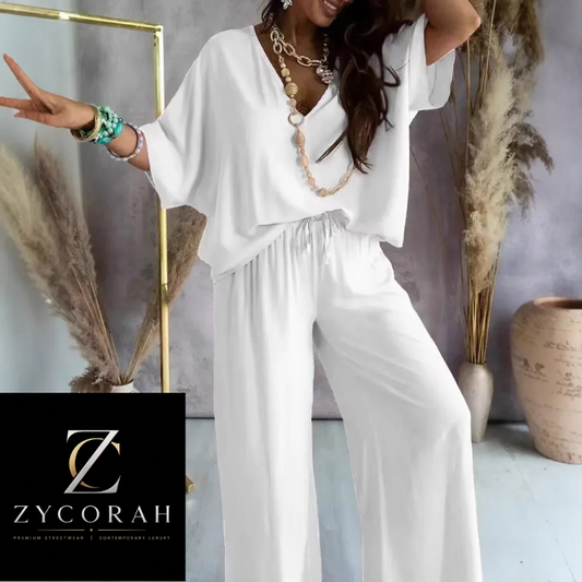 A woman models a white two-piece outfit consisting of a loose short-sleeve V-neck top and matching wide-leg drawstring pants. She is styled with layered gold necklaces and bracelets, standing in a studio setting with dried pampas grass and a ceramic vase.