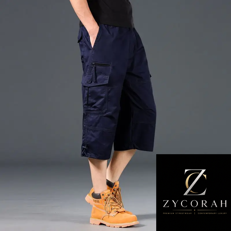 A model wearing dark navy blue cargo shorts with zippered pockets, paired with a black shirt and orange work boots, shown against a gray studio background with a Zycorah brand logo in the bottom right corner.