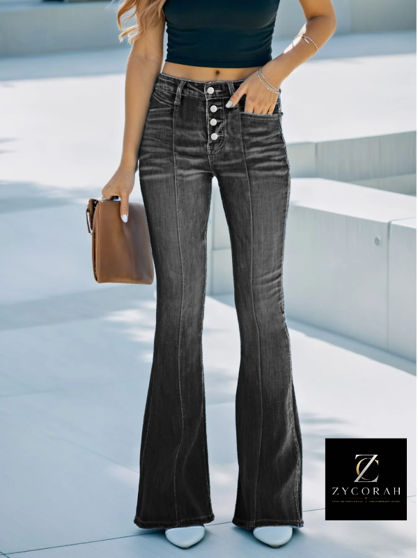 A model wearing high-waisted, dark gray flare jeans with a button fly, standing outdoors on a light concrete surface. She is holding a brown handbag and wearing a black crop top and white shoes.
