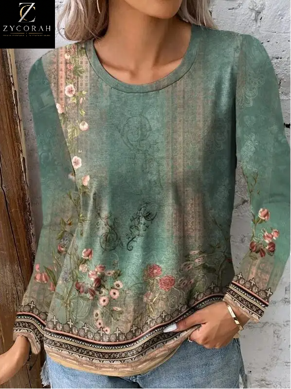 A woman wearing a long-sleeve round-neck blouse with a distressed teal green floral print and a decorative beige patterned hem, shown against a rustic white brick wall.