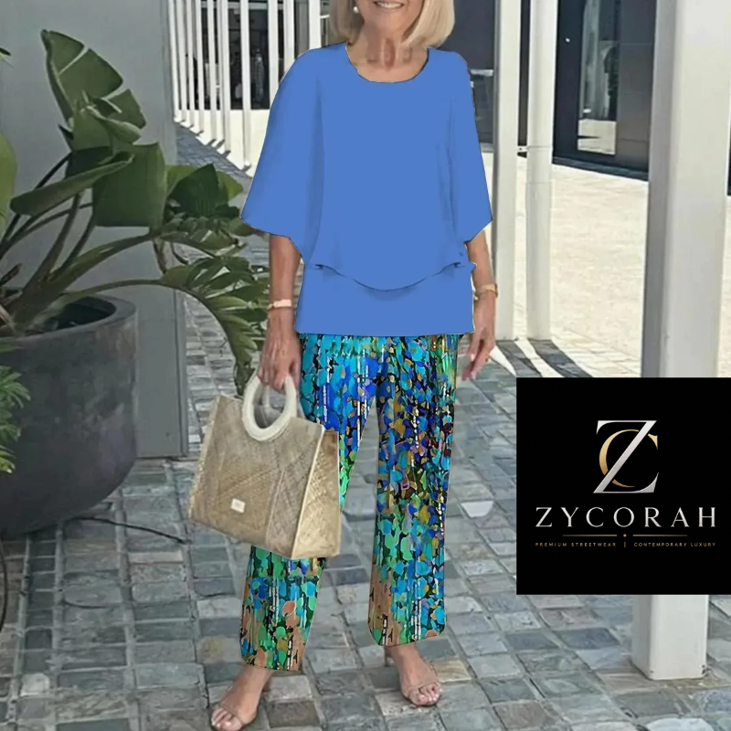 A woman wearing a bright blue short-sleeve top and colorful abstract print pants stands barefoot on a stone walkway, holding a woven tote bag. A ZYCORAH logo overlay appears in the bottom right corner.