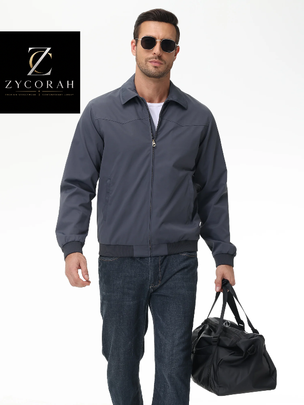 A man wearing a dark slate blue zip-up jacket, dark blue jeans, and sunglasses, holding a black duffel bag. The Zycorah brand logo is visible in the top left corner.
