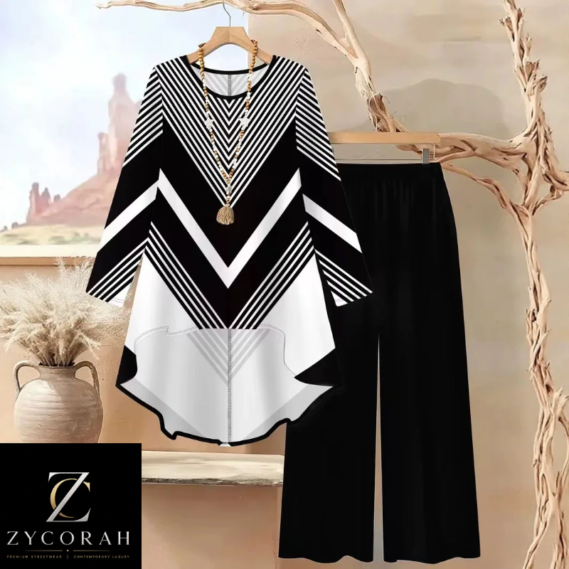 A women's two-piece outfit set featuring a black, white, and gray chevron-patterned long-sleeve top with a high-low hem, paired with solid black wide-leg pants. The set is displayed on a hanger and a wooden hanger respectively against a warm, neutral background with decorative dried branches and a vase.