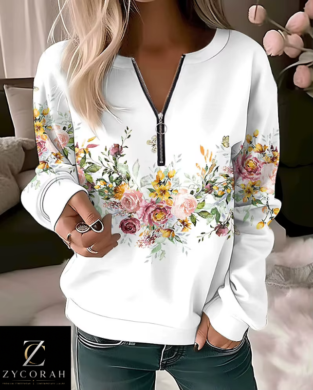 A woman wearing a white long-sleeve half-zip sweatshirt with a colorful floral print across the front and sleeves, paired with dark jeans. The image has a watermark logo for 'ZYCORAH' in the bottom-left corner.