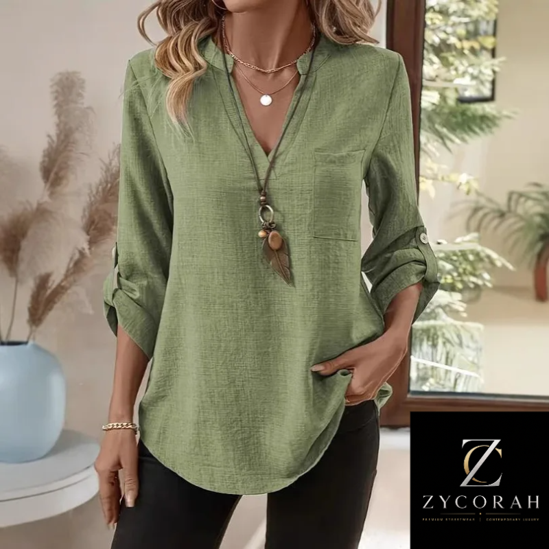 A woman wearing a sage green linen long-sleeve blouse with rolled-up sleeves, a V-neckline, and a chest pocket, paired with black pants. A ZYCORAH brand logo is visible in the bottom right corner.