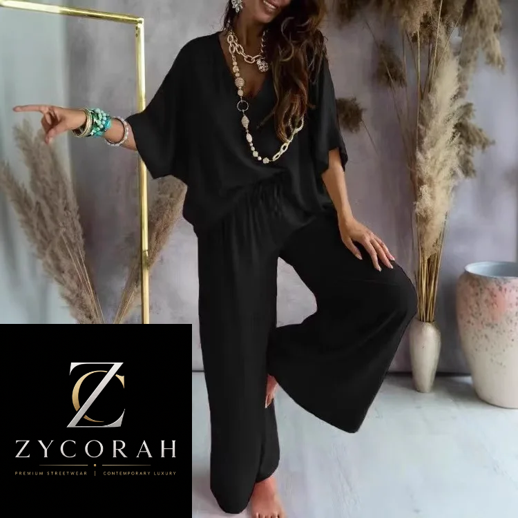 A woman models a black two-piece lounge set consisting of a loose short-sleeve top and wide-leg pants, styled with gold and pearl necklaces and bracelets. The background features a gray wall, a gold mirror frame, dried pampas grass, and ceramic vases.