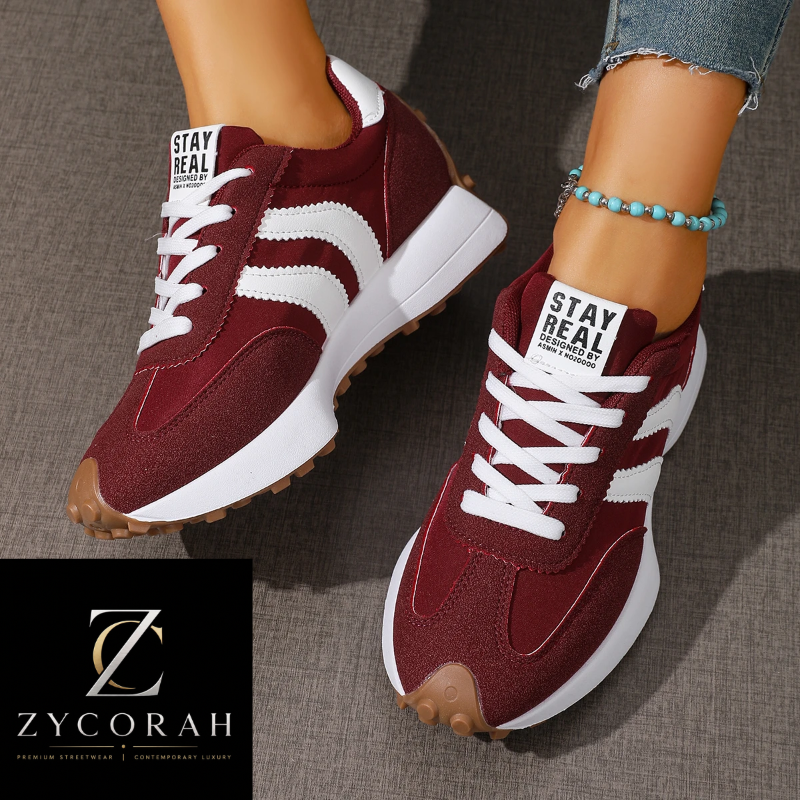 A pair of women's low-top sneakers in burgundy and white, shown being worn on feet with denim jeans visible. The shoes have white laces, white side stripes, a white midsole, and a gum outsole.