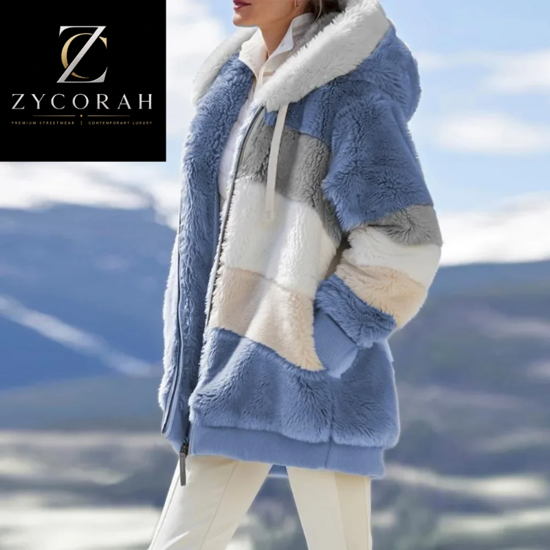 A woman wearing a plush teddy coat with color-blocked panels in light blue, cream, white, and gray, photographed outdoors against a snowy mountain landscape. The Zycorah brand logo appears in the top-left corner.