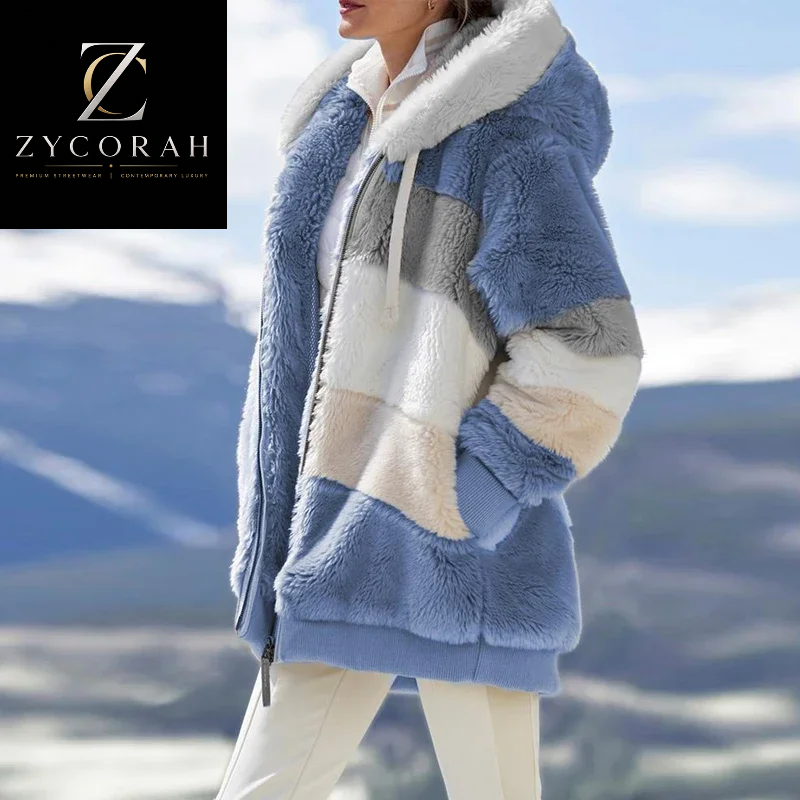 A woman wearing a plush hooded jacket with large color-blocked panels in dusty blue, cream, beige, and light gray, photographed outdoors against a blurred mountain landscape. The Zycorah brand logo appears in the top-left corner.