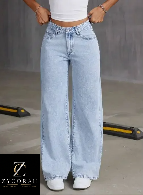 A model wearing light-wash, wide-leg denim jeans with a white crop top and white sneakers, standing in a concrete parking garage. A black brand logo overlay for 'ZYCORAH' is in the bottom-left corner.