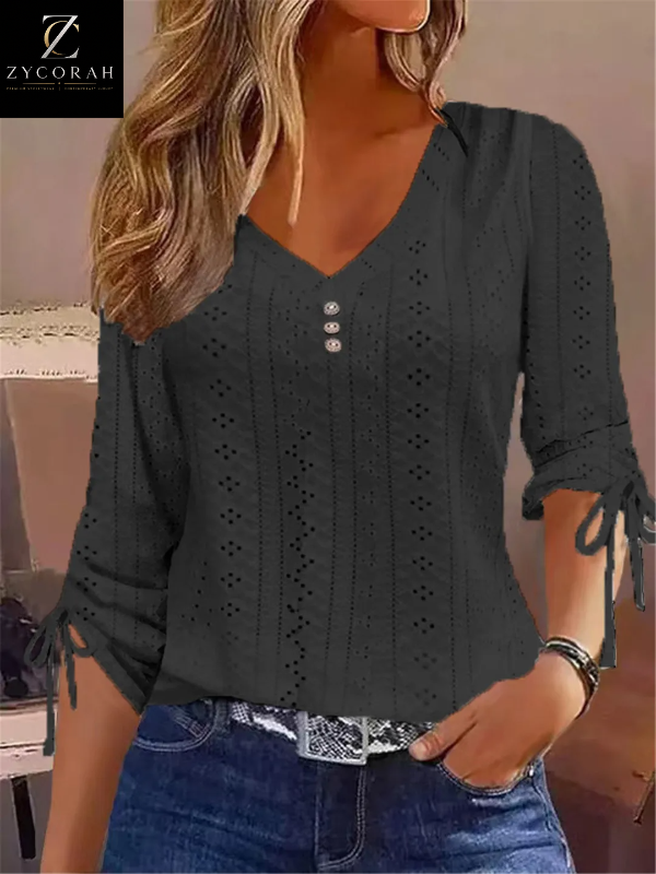 A woman wearing a dark charcoal gray eyelet knit top with a V-neck, three-quarter sleeves, and tie details at the cuffs, paired with blue jeans. The ZYCORAH logo is visible in the top-left corner.
