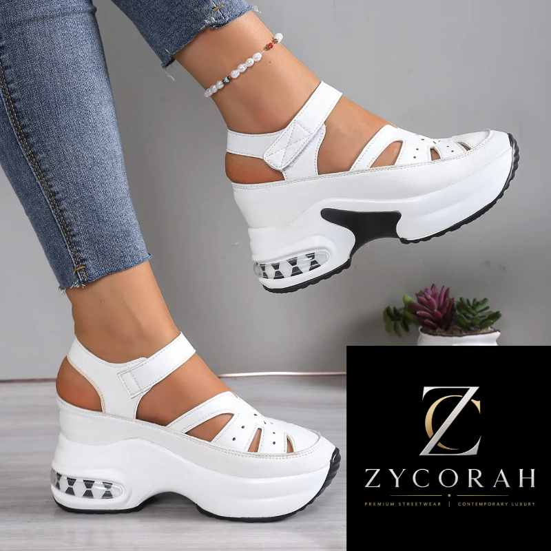 A pair of chunky white platform sandals with cutout straps and ankle straps, shown on a model's feet. The model is wearing blue jeans, and a black brand logo for ZYCORAH is visible in the bottom right corner.