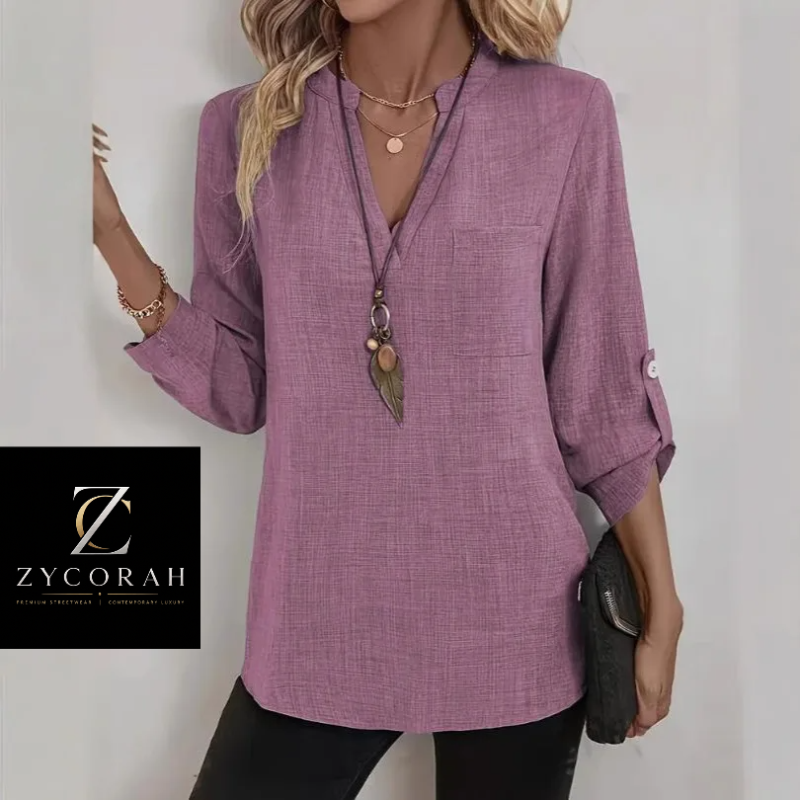A woman models a lavender-purple textured long-sleeve blouse with a split V-neck, chest pocket, and rolled cuffs. She is wearing layered gold necklaces and holding a black clutch.