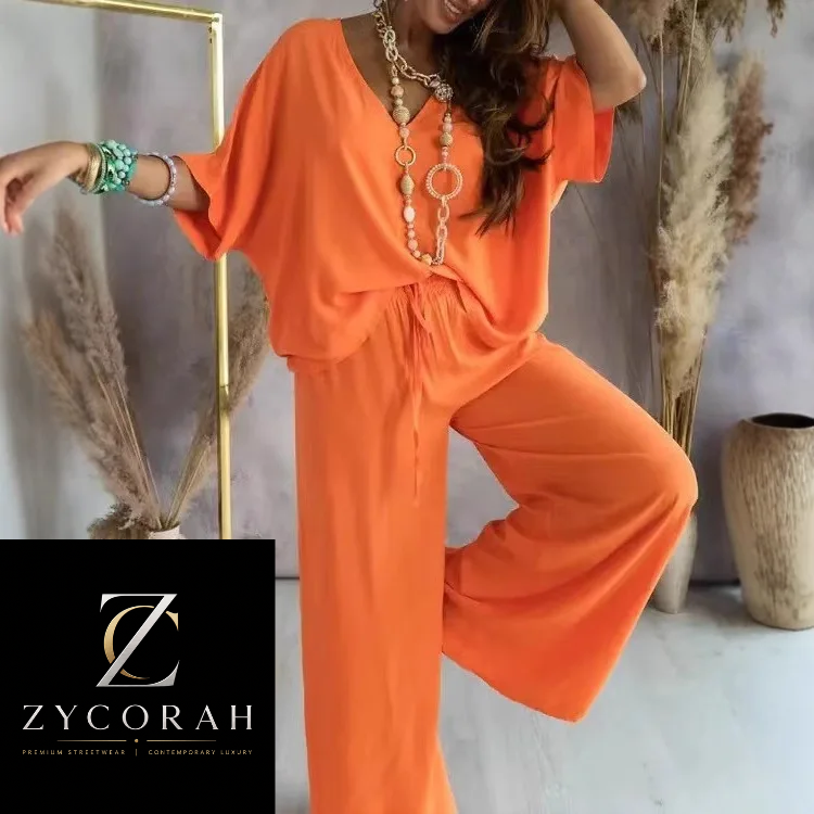 A woman modeling a bright orange two-piece jumpsuit with wide short sleeves and wide-leg pants, styled with layered gold necklaces and bracelets. The background features a light gray wall, a gold-framed mirror, and dried pampas grass in vases.
