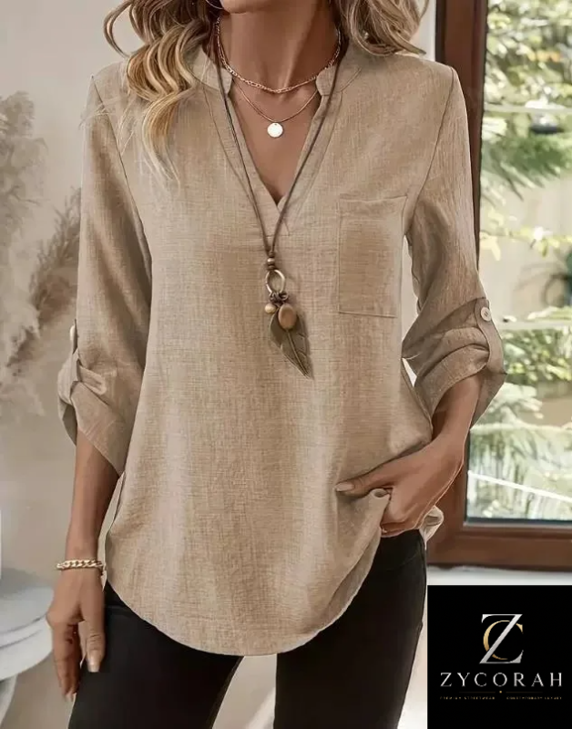 A woman wearing a beige linen-blend blouse with a split V-neck, long sleeves rolled to the forearms, and a chest pocket, paired with black pants. A Zycorah brand logo is visible in the bottom right corner.