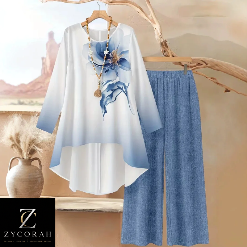 A women's two-piece outfit set featuring a long-sleeve high-low tunic top with a blue floral print and blue gradient hem, paired with matching blue wide-leg pants. The set is displayed on hangers against a warm, desert-themed backdrop with a decorative vase and branch.