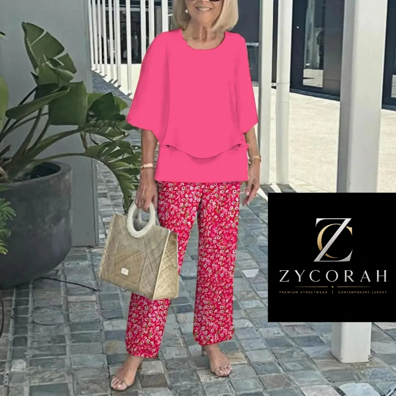 A woman stands barefoot on a stone walkway wearing a bright pink short-sleeve top and red floral pants, holding a woven beige handbag. A black Zycorah logo overlay appears in the lower right corner.