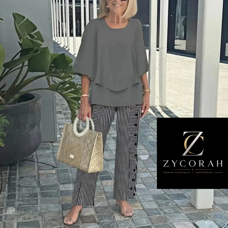 A woman wearing a grey short-sleeve peplum top and black-and-white striped trousers, holding a woven beige handbag. She is standing on a stone walkway next to a potted plant, with a black ZYCORAH brand logo overlay in the bottom right corner.