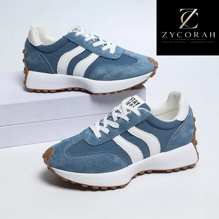 A pair of blue suede and mesh sneakers with white side stripes, white laces, white midsoles, and brown gum outsoles. One shoe is displayed on a white riser and the other lies flat below it.