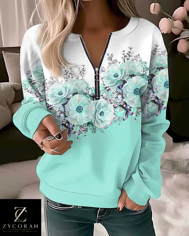 A woman wearing a mint green and white long-sleeve sweatshirt with a floral print across the chest and shoulders, featuring a half-zip neckline. The sweatshirt has a solid mint green lower half and a white upper half with a large floral pattern in shades of teal, blue, and purple.