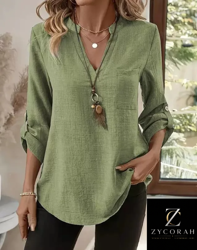 A woman wearing a sage green linen long-sleeve blouse with a split neckline, chest pocket, and rolled sleeves, paired with black pants. The Zycorah brand logo is visible in the bottom right corner.