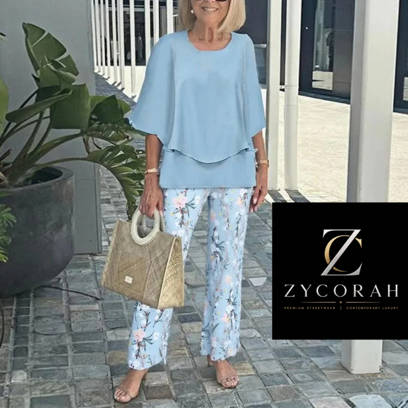 An older woman stands outdoors wearing a light blue flutter-sleeve top and light blue floral pants, holding a woven beige handbag. A black brand box in the lower right reads ZYCORAH.