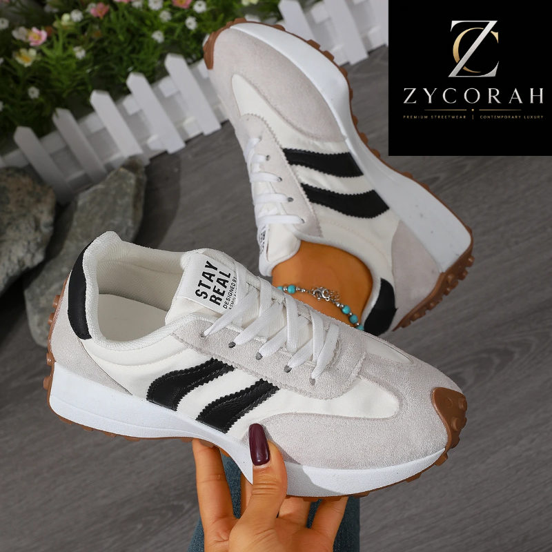 A pair of low-top sneakers held in a hand against a gray floor background, with a white picket fence and flowers in the upper left. A black Zycorah logo overlay appears in the top right corner.