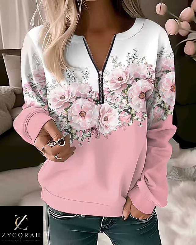 A woman wearing a pink and white long-sleeve sweatshirt with a floral print across the chest and shoulders, paired with dark teal jeans. The sweatshirt features a half-zip neckline with a black zipper.