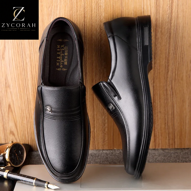 A pair of black leather slip-on loafers shown on a tabletop against a wood panel background, with a ZYCORAH brand logo overlay in the top-left corner and a watch and pen as styled props.