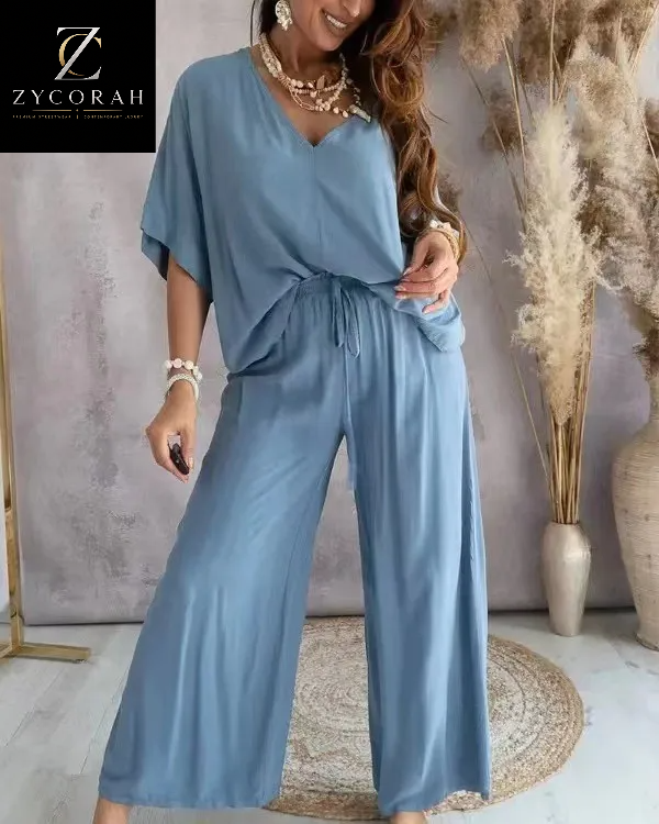 A woman models a light blue two-piece jumpsuit featuring a V-neck top with short wide sleeves and matching wide-leg pants. The image includes a 'ZYCORAH' brand logo in the top-left corner, set against a styled indoor background with dried pampas grass in a vase.