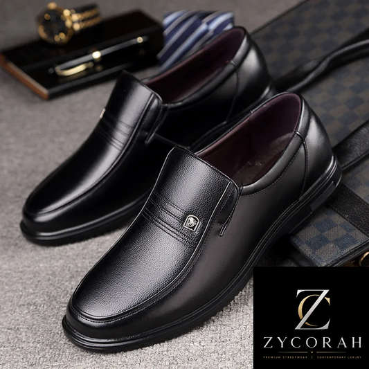 A pair of black leather slip-on loafers with a small metal emblem on the vamp, displayed on a gray textured surface with a striped tie, watch, and patterned bag as styling props. A ZYCORAH brand logo overlay is in the bottom right corner.