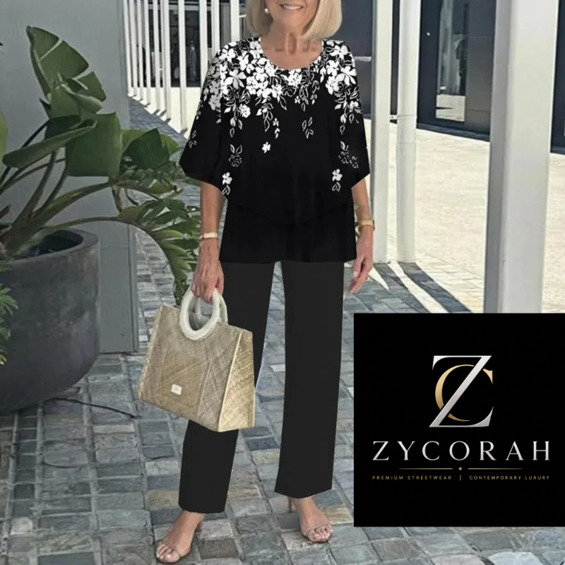 A woman wearing a black short-sleeve blouse with white floral embroidery and black wide-leg pants, holding a woven beige handbag. She is standing outdoors on a stone walkway next to a large potted plant.