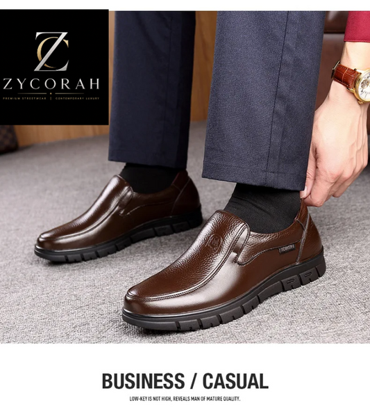 A man wearing dark brown slip-on loafers with a textured grain upper and black soles, paired with navy trousers and black socks. The ZYCORAH brand logo is visible in the top left corner, and the text 'BUSINESS / CASUAL' is at the bottom.
