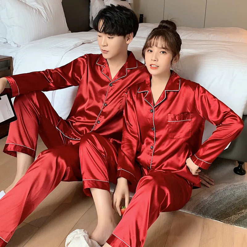 A man and a woman wearing matching shiny red satin pajama sets with white piping, sitting together in a bedroom with a bed and wooden floor visible.